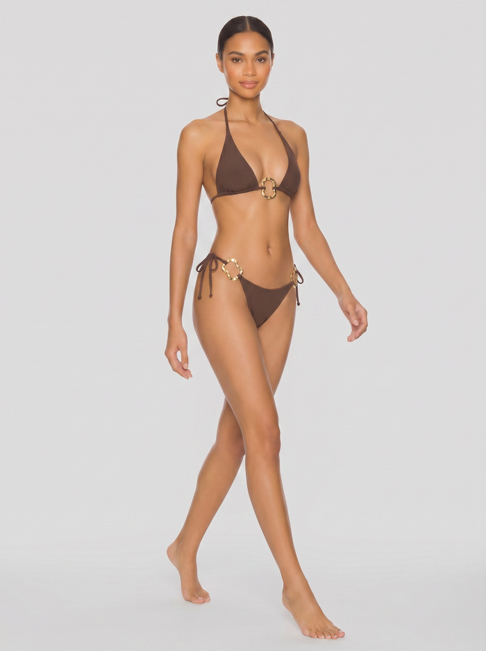 Brown triangle bikini with gold ring accents, side-tie bottoms, model walking against a light gray studio backdrop